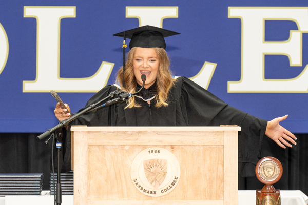Student Rosie Luke wearing graduation regalia while standing at podium spreading her arms wide in celebration