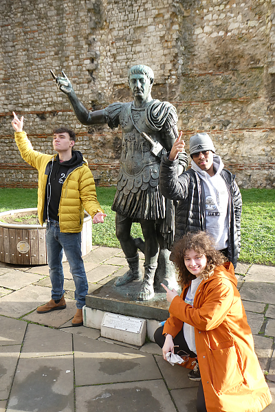 3 studeents pose with statute. Two are mimicking the statue's pose. The third is crouched below and pointing at it.