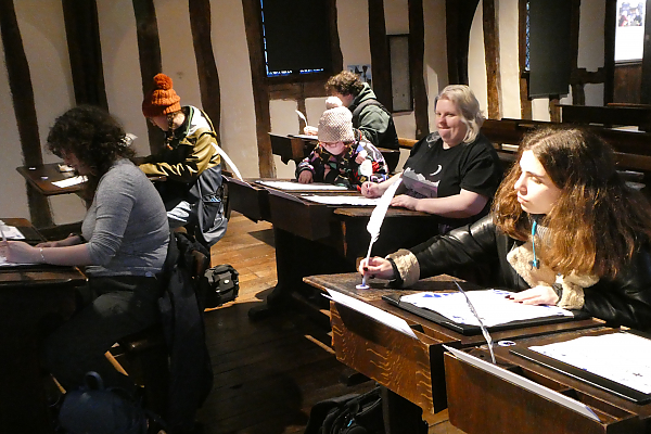 Six students sitting at wooden desks writing with quill pens