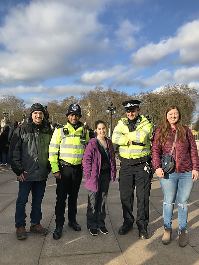 From left to right, a male student, a police officer, a female student, a police officer, and a female student smile for the camera. Both officers are wearing bright yellow reflective vests over their uniforms.