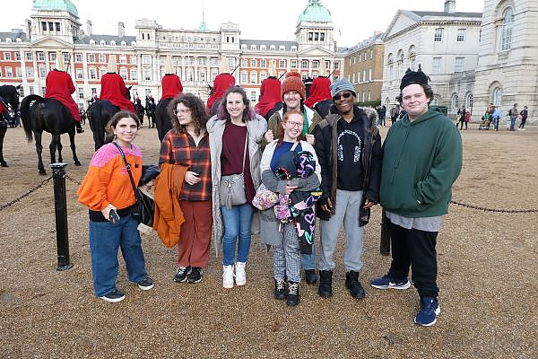 Seven students posing for a photo with men on horses lined up behind them and Buckingham Palace in the far background