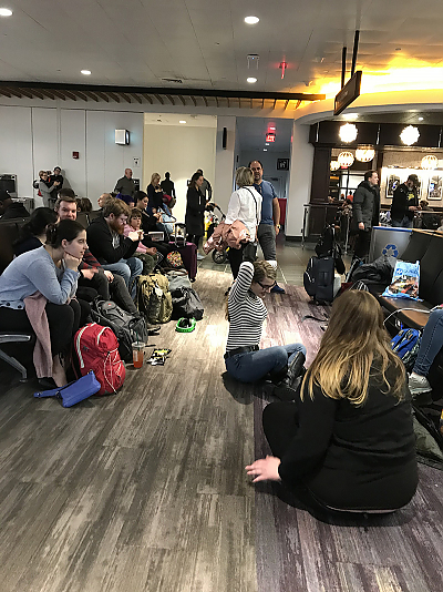 A shot of the Logan Airport terminal with student seated in chairs or on the floor.