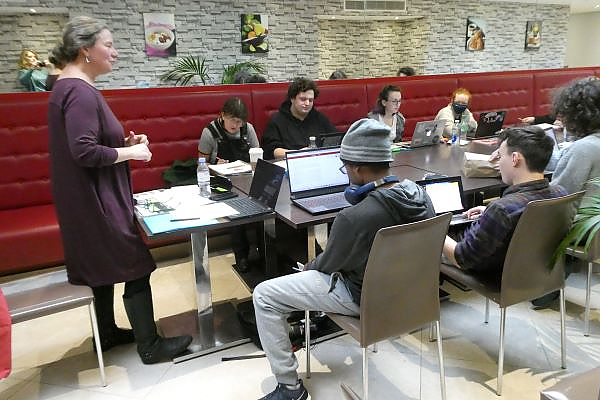 Students sitting at a long table with laptops open. At the head of the table, Professor Adrienne Major is standing and addressing the students.