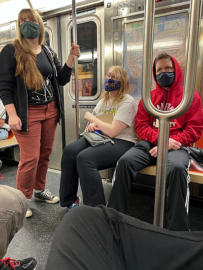 Three students, one standing and two seated, on a subway car looking past the camera at