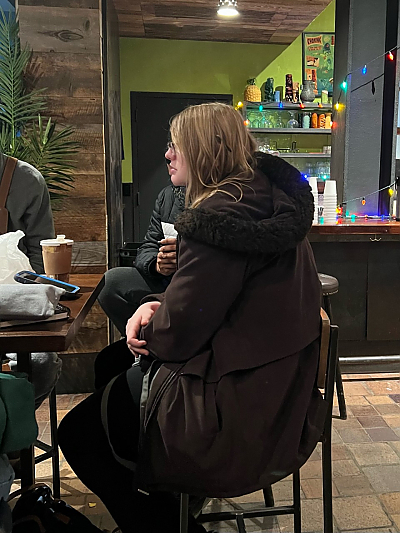 Profile of student with shoulder length blond hair and glasses seated at table listening to discussion. They are wearing a long brown winter coat and have their arms folded.