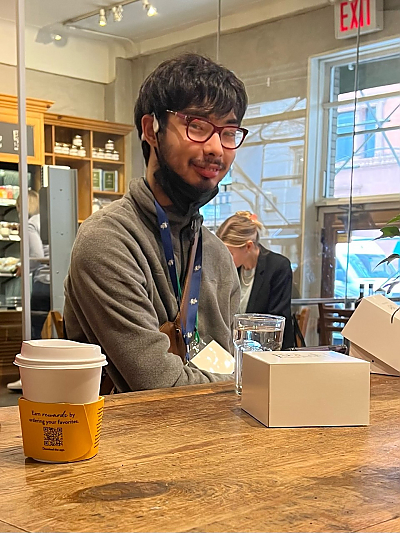 A student with dark hair, glasses and mask pulled down smiles at camera while sitting at a cafe table with glass of water and coffee cup on it.