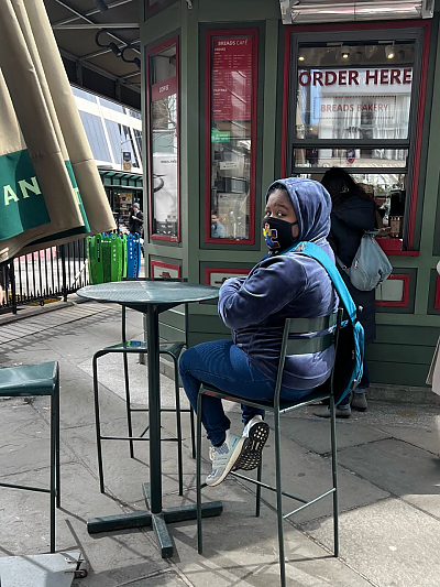 Student looking over her shoulder at camera while sitting at outdoor cafe table with purple hoodie pulled up over her head and wearing protective mask