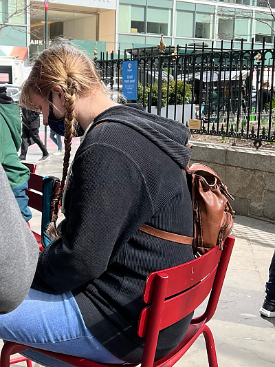 Over the shoulder photo of female student with long blond braids sitting outdoors in red chair while looking downward at something not visible