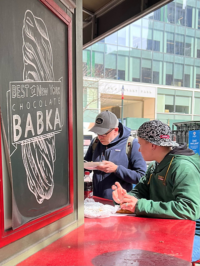 Two people wearing baseball caps seated outdoors eating lunch and talking to each other. in the foreground is a chalkboard sketch of a pastry and text that reads Best of New York chocolate babka