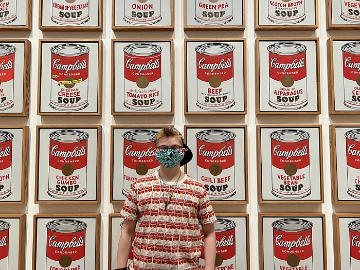 Student standing in front of Andy Warhol soup can painting while wearing a shirt with a print of the same piece of art.
