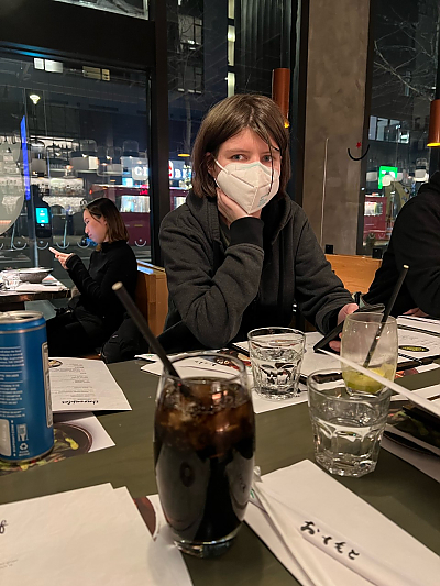 Student wearing protective mask sitting at table holding smart phone but looking up past camera
