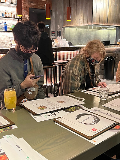 2 students wearing protective masks, sitting in restaurant. Male student on left is looking at his smartphone. Female student on right is looking at the menu.