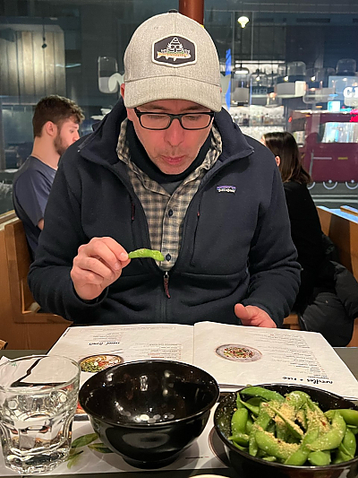 Man in baseball cap holding piece of edamame while reading menu at New York restaurant