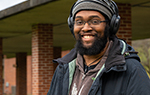 Photo of Anastasio Bonhomme smiling for camera on Landmark College quad. He is wearing a wool hat and headphones. 