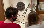 An over the shoulder picture of two students looking at an art exhibit of small disc and kniives.