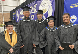 Group of five students standing on graduation stage wearing caps and gowns posing for a photo before the December 17 Commencement ceremony