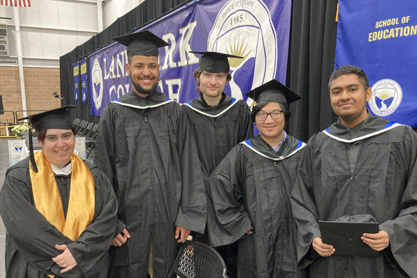 Group of five students standing on graduation stage wearing caps and gowns posing for a photo before the December 17 Commencement ceremony