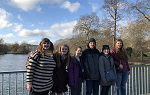 Six students pose for a picture outdoors on a bridge on a sunny day. Their backs are leaning against the railing with a river behind them. Their clothing indicates the weather is chilly.  