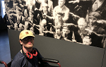 Student in wheelchair with glasses and yellow baseball cap poses for picture in front of a large black and white image in the German Resistance Memorial Center.  