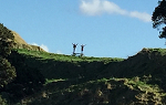 A photo of from the 2018 Summer Study Abroad trip to New Zealand, showing two students standing on an outdoor picnic table with their arms raised. The photo is taken from a far distance, showing the grandeur of Mount Maunganui.  