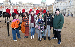 Seven students posing for a photo with men on horses lined up behind them and Buckingham Palace in the far background
