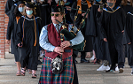 A man playing bagpipes and wearing kilt leads students wearing graduation caps and gowns toward commencement area