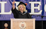 A woman with blond hair wearing black graduation cap and gown stands at podium giving the peace sign
