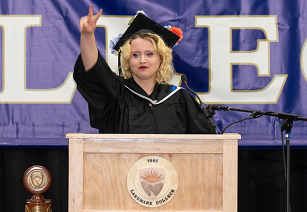 A woman with blond hair wearing black graduation cap and gown stands at podium giving the peace sign