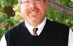Headshot of Dr. David Parker. He is facing the camera and smiling. He is wearing a dark colored sleeveless sweater with a white shirt and necktie. He has reddish-orange hair with a graying goatee and glasses. 