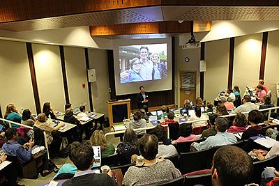 Brooks M. O'Brien auditorium full of participants watching presenter. Image of people on screen at front of room.