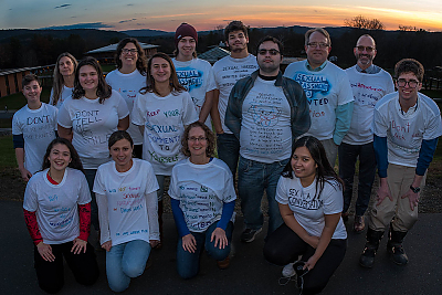 A group of roughly a dozen students, faculty and staff wearing t-shirts made for Sexual Harassment Awareness Day. They are standing outside on the upper campus with an evening view of the mountain range behind them. 