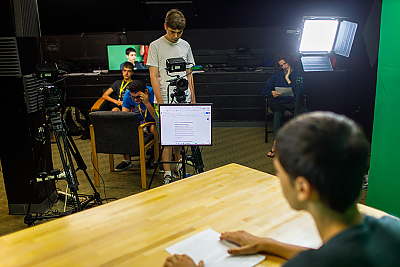 Over the shoulder image of student at a table. He is facing a student operating a camera with a teleprompter in front of it. 