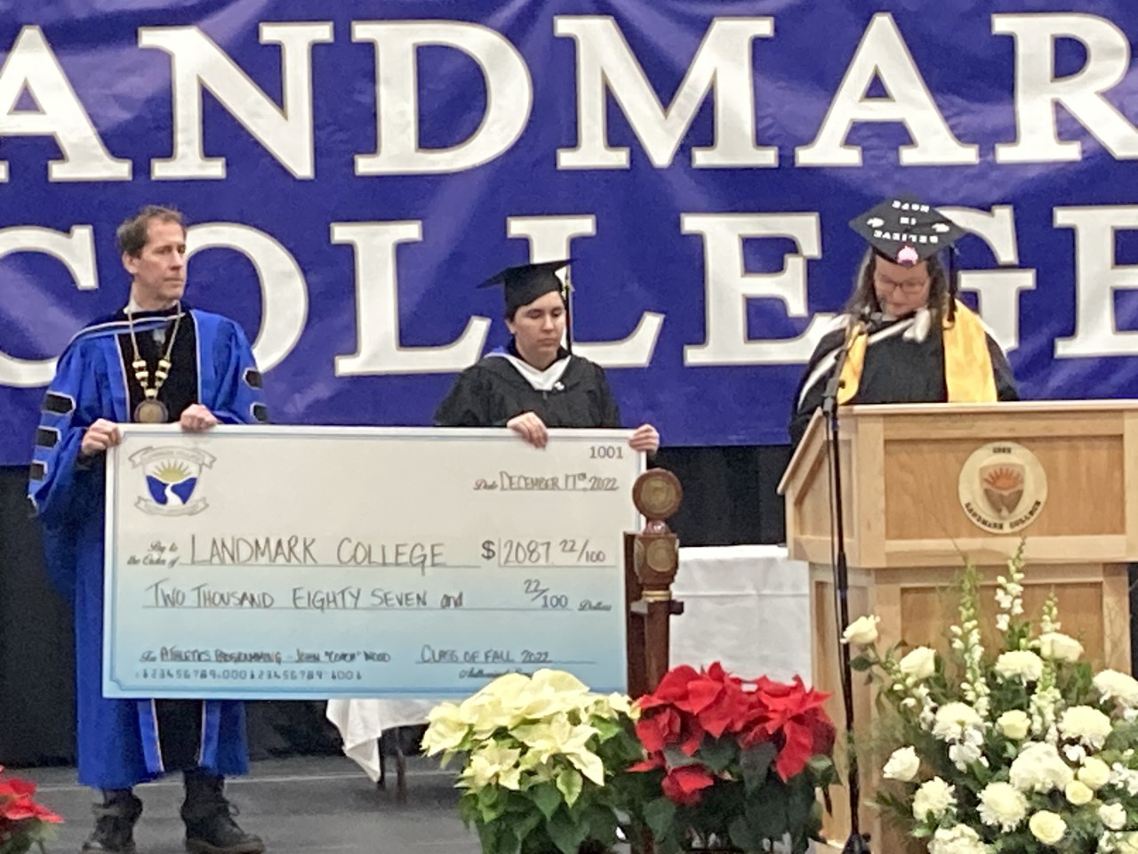 Image of President Eden and graduating student holding oversized check in the amount of $2,087.22, while another graduating student makes remarks at the podium.