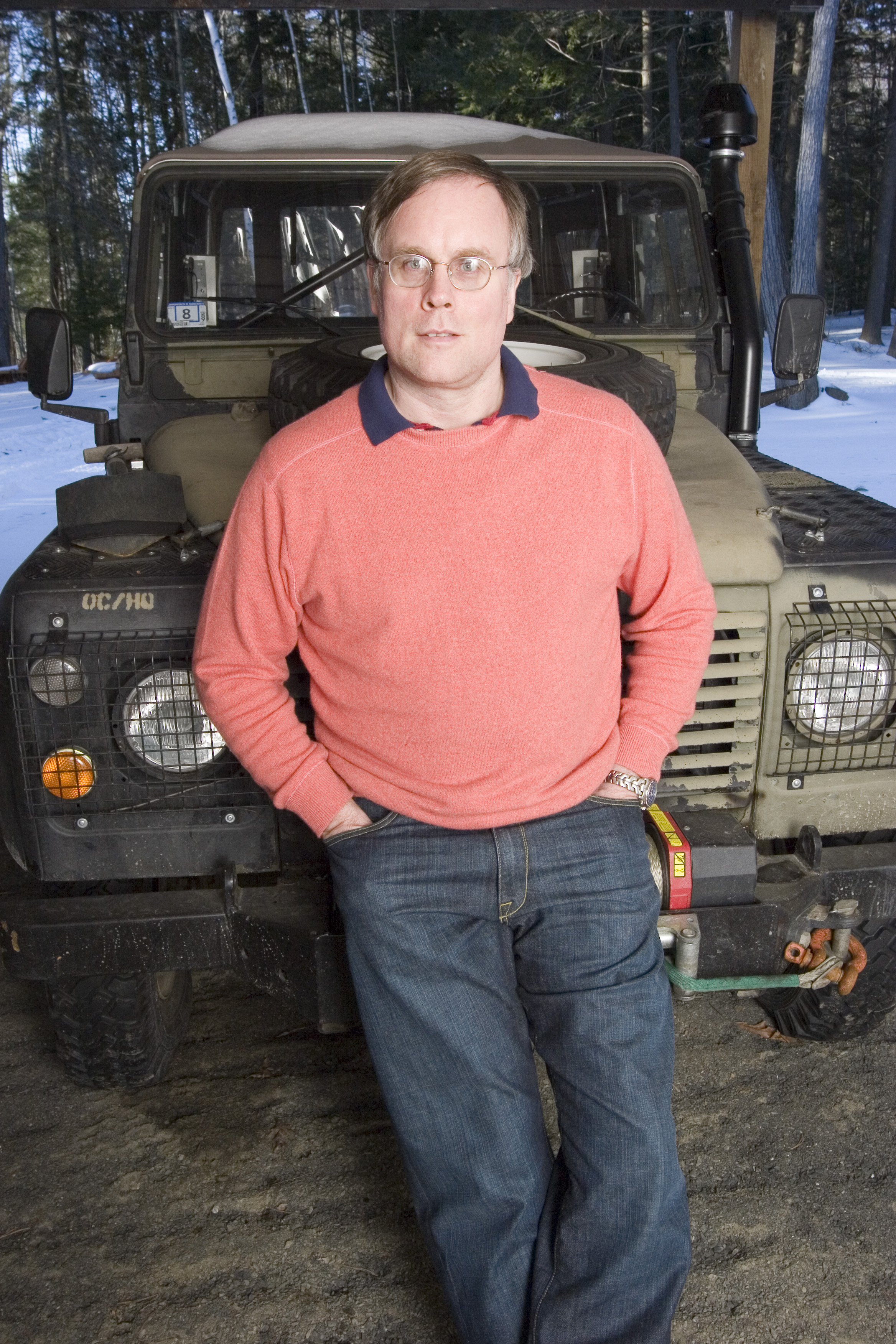 Photo of man wearing orange sweater and glasses posing for photo while leaning on the hood of a jeep. 