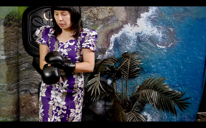 A woman with dark hair and a flower print dress standing in front of a screen showing an overhead view of an ocean shore. She is putting on boxing gloves. 