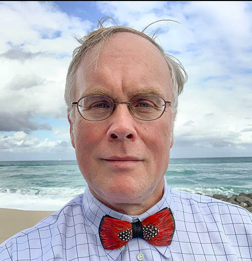Headshot of John Elder Robison outside with an ocean backdrop. He is wearing a blue checked shirt, red bowtie and glasses