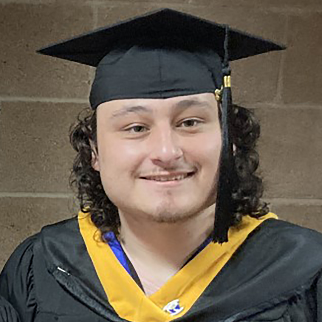 Young man with long curly black hair and slight goatee smiles at camera. He is wearing a graduation cap and gown. 