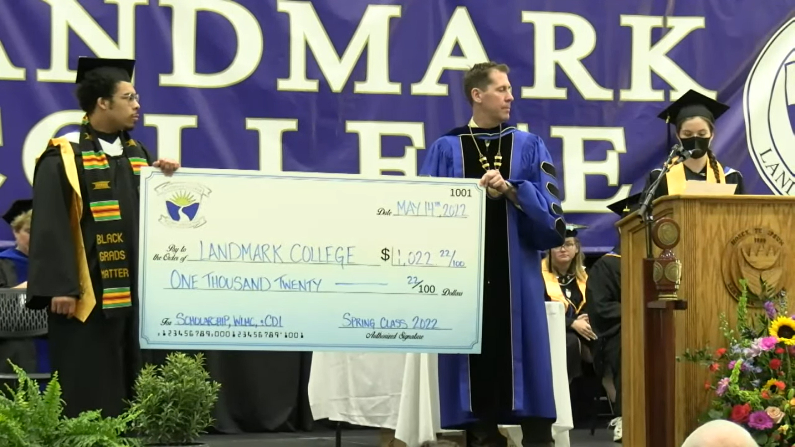 Image of student Carter Griffith and President Peter Eden, both in graduation regalia, standing on the commencement stage holding an oversized check in the amount of 1,022.22. They are both looking to their left  at student Meredith Robertson, who is making remarks at the podium while wearing a protective mask.