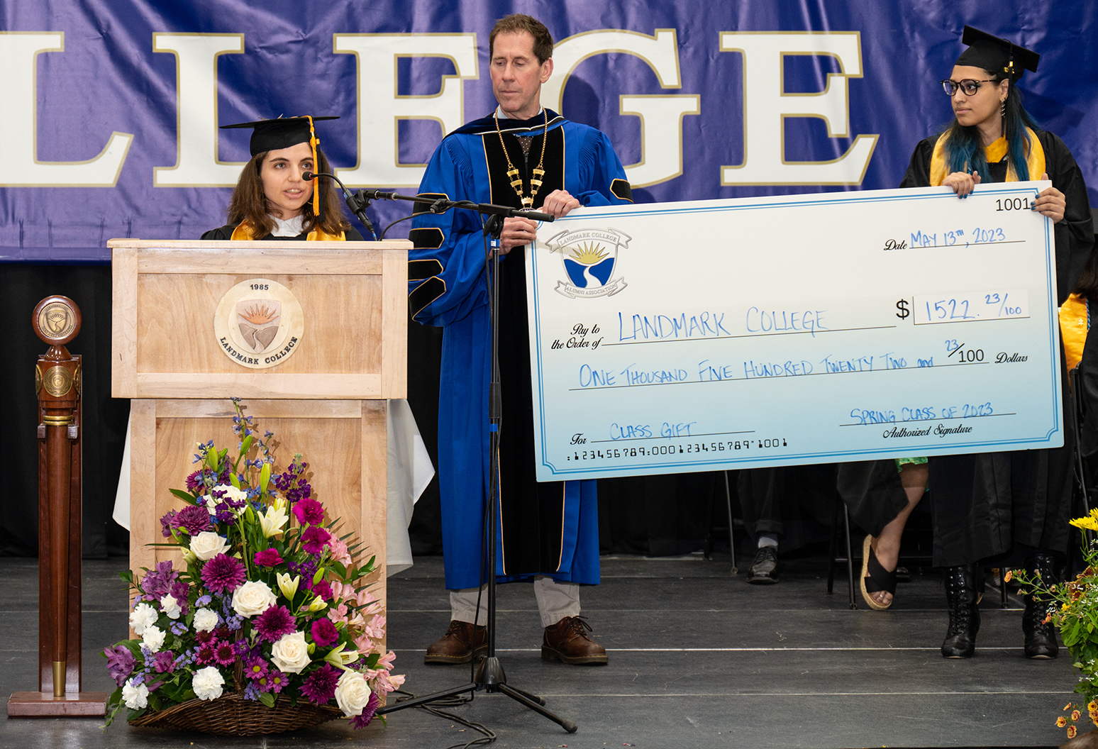 A female student stands at podium making remarks while a male and female hold an oversized check made out to Landmark College in the amount of $1,522.23 