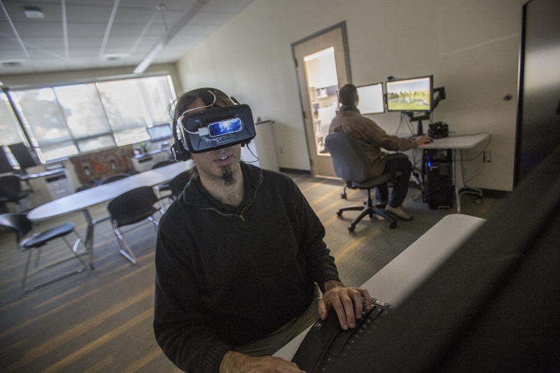 Researcher wears virtual reality headset and faces camera while other researcher works at computer monitor in background