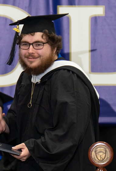 Man with bushy beard and dark rimmed glasses wearing black graduation cap and gown smiles while shaking hands with someone off camera