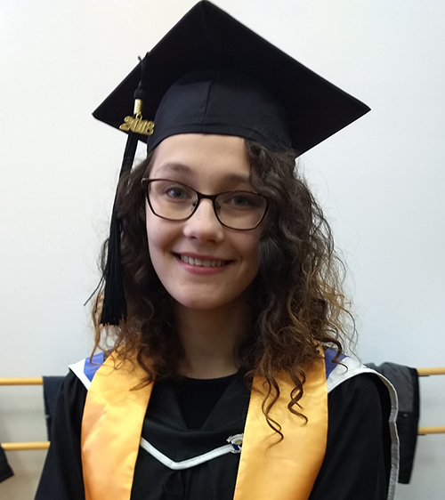 Katie LaBombard, a woman with long, curly brown hair and glasses. She is dressed in a black graduation cap and gown with a gold honors sash