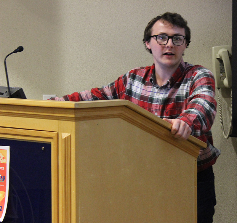 Male student with glasses and checked shirt making remarks while standing at a podium