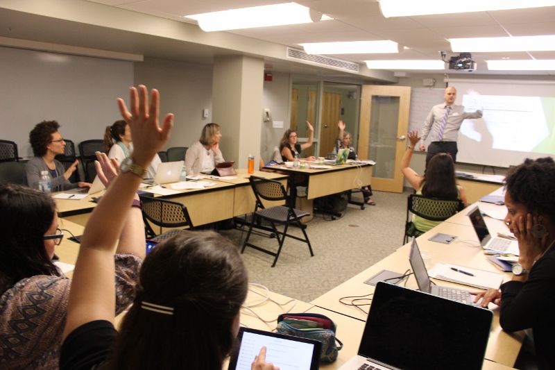 Participant in foreground raises hand while LCIRT presenter speaks to students around tables in Landmark College classroom