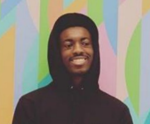 Julius Udochi standing in front of a colorful background, wearing a black sweatshirt with the hood pulled up